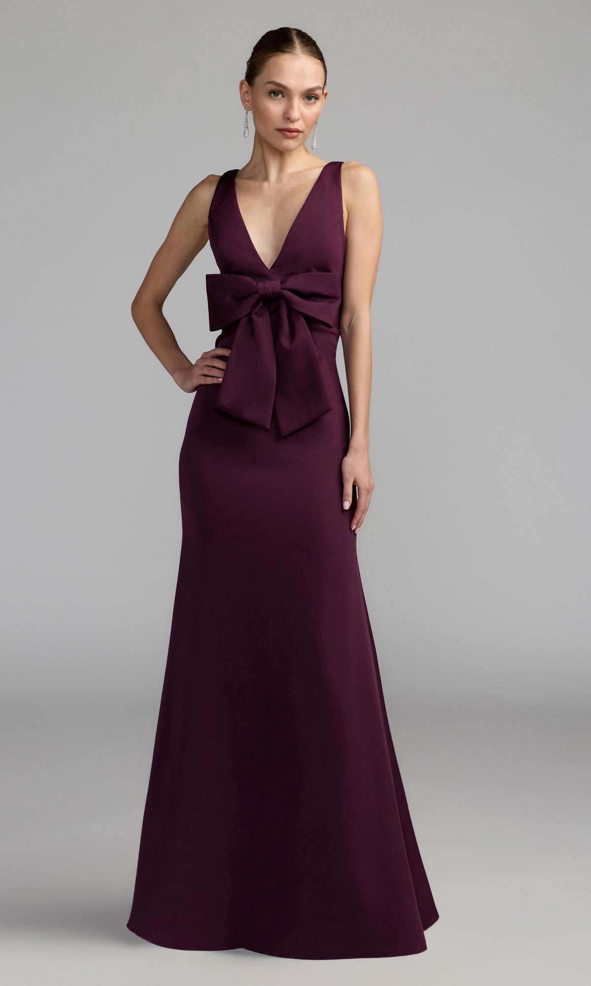 Woman wearing a sleeveless, floor-length Mac Duggal 50087 Bow-Front Green Formal Dress with a deep V-neckline and large waist bow, standing against a plain gray background.
